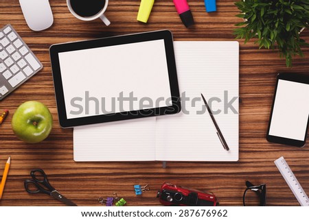 Modern black digital tablet with blank screen on notebook in the middle of office equipment such as computer keyboard, mouse, cup of coffee, smartphone, apple pencil  on dark wooden office desk.