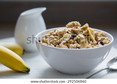 Bowl of muesli with dried banana, spoon, pitcher of milk, fresh banana on white pad.