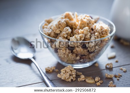 Bowl of muesli with dried banana, spoon, pitcher of milk on wooden table with banana and muesli pieces scattered all around.