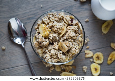 Top view on bowl of muesli with dried banana, spoon, pitcher of milk on wooden table with banana and muesli pieces scattered all around.