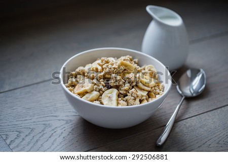 Bowl of muesli with dried banana, spoon, pitcher of milk and spoon on wooden table.