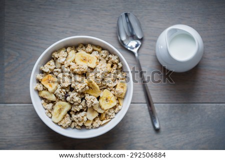 Top view on bowl of muesli with dried banana, spoon, pitcher of milk and spoon on wooden table.