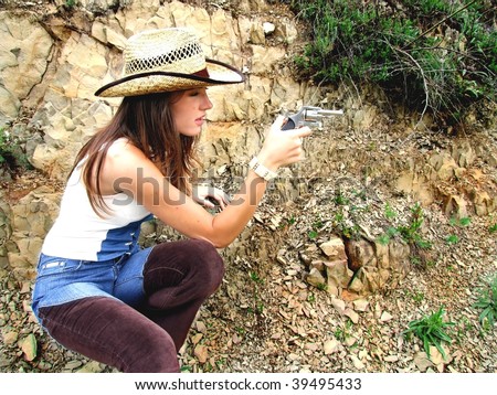 Young adult female poses in a crouch wearing a western style straw hat and aiming a small pistol.  Rugged canyon is the backdrop.