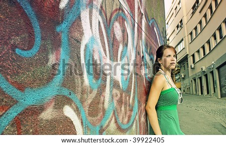 Angled profile shot of a teenage girl leaning against a graffiti wall in the city, with her face turned toward the viewer.