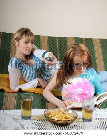 Two beauty girl friends, reading book and watching TV with juice and some snacks on table.