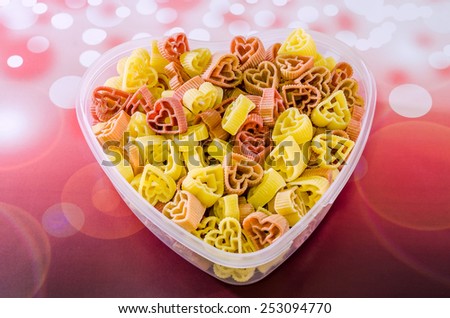 Transparent heart shape vase (bowl) filled with colored (red and yellow) heart shape pasta, red bokeh background, close up