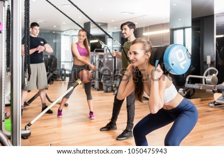 Young determined fit woman doing squats exercise, while holding on the back of her neck a heavy sandbag during group circuit functional training at the gym