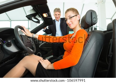 Young woman with mature man sits in car on driver seat with her hand on the steering wheel in a car dealership, obviously she is buying or selling the convertible