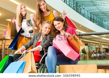 Four female friends with shopping bags having fun while shopping in a mall, stores in the background; one woman is sitting in a wheelchair