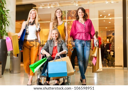 Four female friends with shopping bags having fun while shopping in a mall, stores in the background; one woman is sitting in a wheelchair