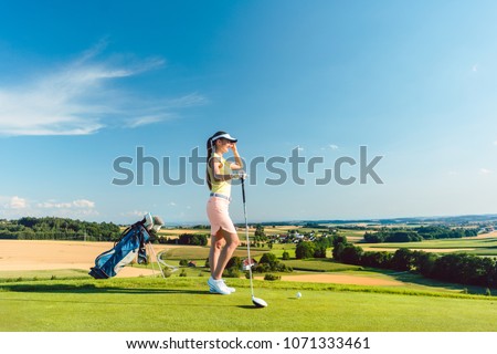 Side view of a fit woman wearing modern golf outfits, while looking at the horizon on the green grass of a golf course in the countryside in a sunny day of summer