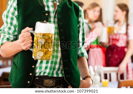 Young people in traditional Bavarian Tracht in restaurant or pub, one man is standing with beer stein in front, the group in the background