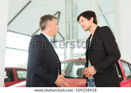 Mature single man with red auto in light car dealership with a male customer, a young man, he is obviously buying a car or is a car dealer
