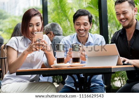 Three young Asian friends smiling while using electronic devices connected to the wireless internet network of a modern coffee shop