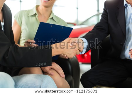 Two men in business suits shaking hands after a successful car purchase in front of a woman holding documents in a light car dealership