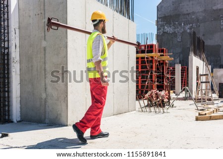 Full length of a blue-collar worker wearing safety equipment, while carrying a heavy metallic bar during work on the construction site of a residential building