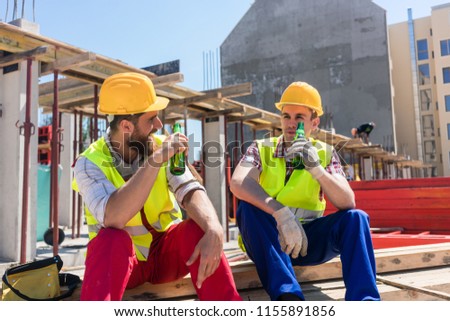 Two young workers smiling while drinking a cold alcoholic or non-alcoholic beer, during break at work on the construction site in a sunny day of summer