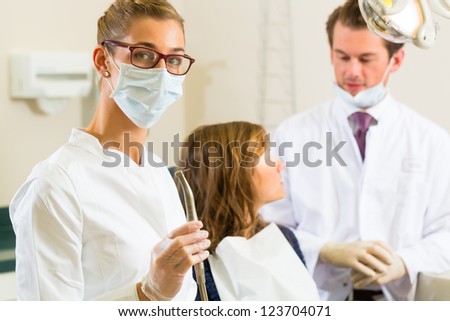 Dentists in his surgery holds a drill and looking at the viewer, in the background her colleague is giving a female patient a treatment