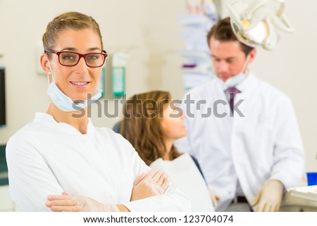 Dentists in her surgery looking at the viewer, in the background her colleague is giving a female patient a treatment