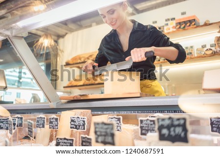 Young shop clerk in deli cutting cheese at the counter with a knife