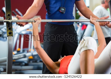 Woman with her personal fitness trainer in the gym exercising with dumbbells, she is using barbell on a weight bench