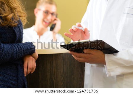 Patient and doctor in reception area of office of doctor or dentist, he holds a clipboard, the receptionist is on the phone in background