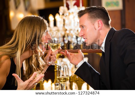 Couple have a quarrel on a romantic date in a fine dining restaurant they are angry and yelling, a large chandelier is in Background