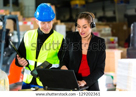 Teamwork - warehouseman or forklift driver and female supervisor with laptop, headset and cell phone, at warehouse of freight forwarding company - a forklift is in Background