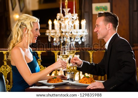 happy couple have a romantic date in a fine dining restaurant they drink wine and clinking glasses, cheers - a large chandelier is in Background