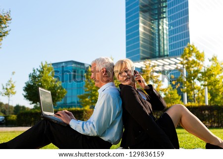 Business people working outdoors - he is working with laptop, she is calling someone on phone