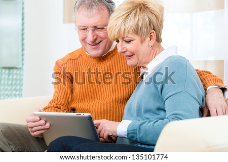 Quality of life - two elderly people sitting at home on the couch and writing emails on the tablet computer