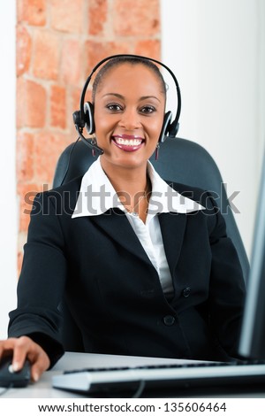 Young businesswoman or secretary working in her Office, she is sitting on the desk in front of the window and working on a computer with a headset, she has a customer pitch