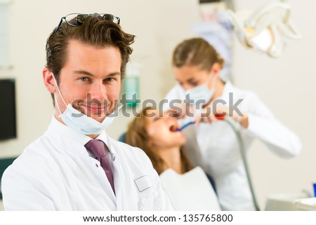 Dentists in his surgery looking at the viewer, in the background his assistant is giving a female patient a treatment
