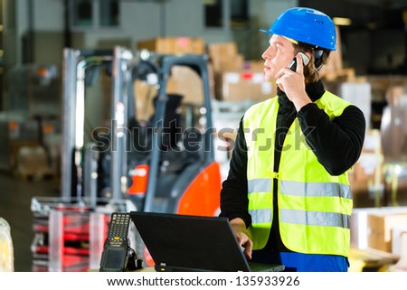 Warehouseman with protective vest, scanner and laptop in warehouse at freight forwarding company using a mobile phone
