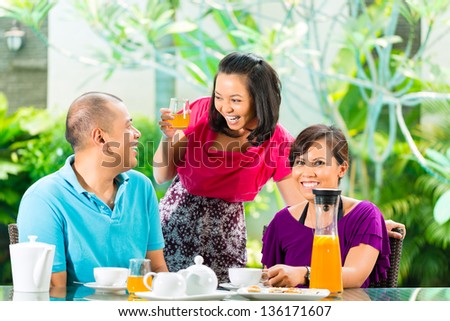 Asian friends having coffee on the porch in front of a home, in the background a tropical garden