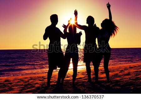 People (Two Couples) On The Beach Having A Party, Drinking And Having A Lot Of Fun In The Sunset (Only Silhouette Of People To Be Seen, People Having Bottles In Hands With The Sun Shining Through)