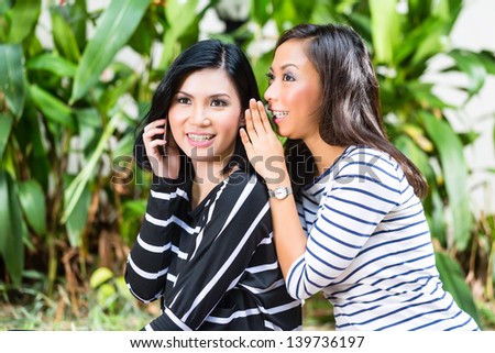 Two Indonesian friends, in a tropical environment, one with Smartphone, the other whispers to her, and gives tips
