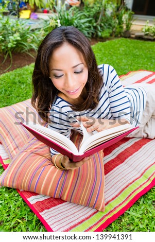 Young Indonesian woman lying at home in the garden and reading a book in her leisure time