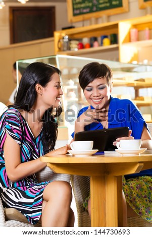 Asian female friends enjoying her leisure time in a cafe, drinking coffee or cappuccino and looking at photos or emails on a tablet computer