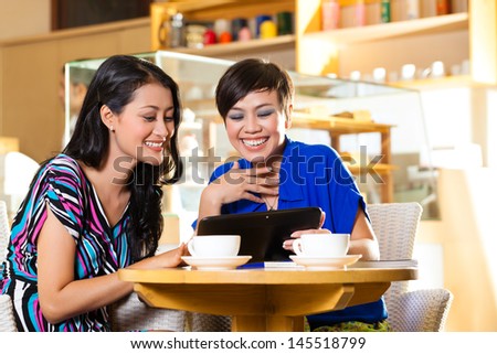 Asian female friends enjoying her leisure time in a cafe, drinking coffee or cappuccino and looking at photos or emails on a tablet computer
