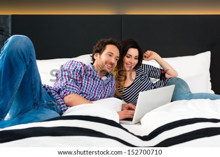 Young Couple lying in the bed of a hotel room, they are on vacation and using the wifi in the room for internet with the computer