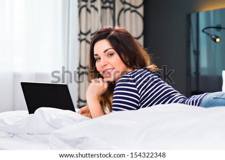 Young woman lying in the bed of a hotel room, she are on vacation and using the wifi in the room for internet with the computer