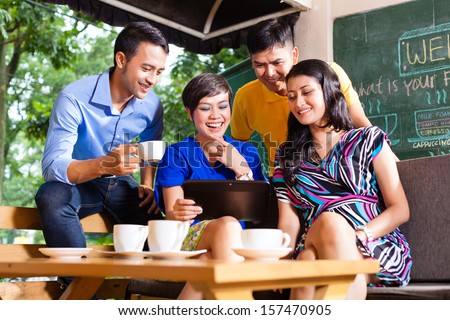 Asian Friends Or Colleagues Enjoying Leisure Time In A Cafe, Drinking Coffee Or Cappuccino And Looking At Photos Or Emails On A Tablet Computer
