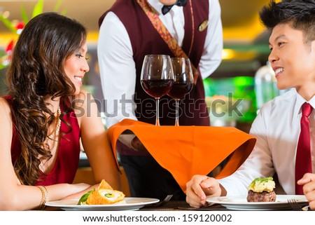 Asian Chinese waiter or steward serving man and woman or couple red wine in glasses on a tray in fancy restaurant or hotel