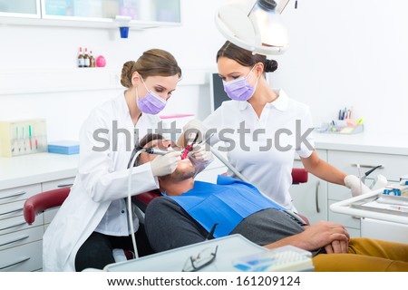 Dentist In Her Practice Or Office Treating Male Patient With Assistant Wearing Masks And Gloves