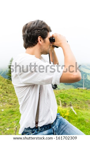 Hiking - Young man in the Bavarian Alps looking through a field glasses or binocular enjoying the panorama in the leisure time or vacation
