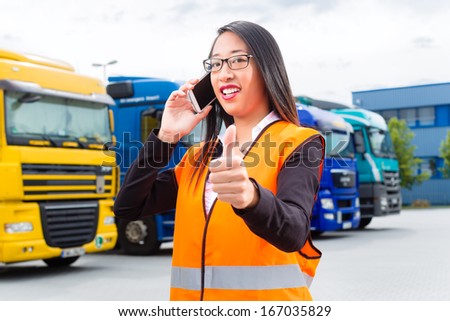 Logistics - female Asian forwarder or supervisor with mobile phone, in front of trucks and trailers, on transshipment point