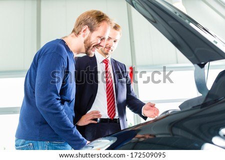 Seller or car salesman and client or customer in car dealership presenting the engine performance of new and used cars in the showroom, the men looking under the hood