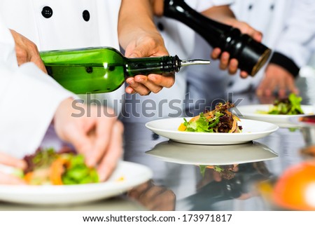 Asian Indonesian Chef Along With Other Cooks In Restaurant Or Hotel Commercial Kitchen Cooking, Finishing Dish Or Plate