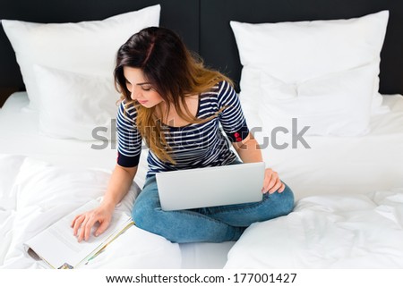 Young woman sitting on bed of a hotel room, she is on vacation and using the wifi in the room for internet with the computer